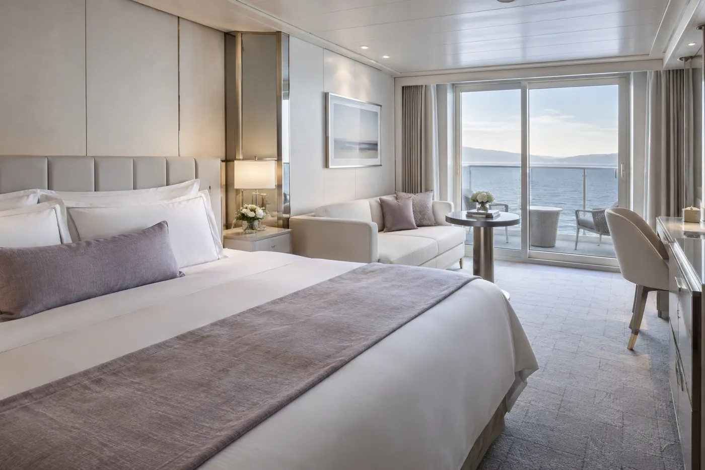 Oyster-white shipboard stateroom with lavender textiles, a sitting area and a private balcony facing the sea